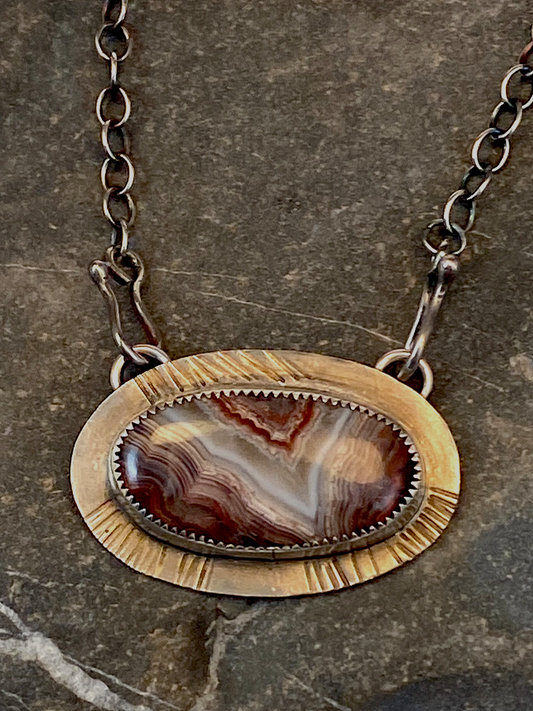 Close up view. Crazy lace agate with beautiful red, white and gray colors graces this one-of-a-kind necklace.   Set on textured  backplate of sterling silver.  19" sterling silver chain with unique handcrafted clasps that attach in front. Approx. 1” tall 2” wide.  Style: rustic, boho, funky, organic, earthy, natural